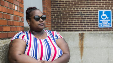 Cynthia Norman stands against a brick wall, sunglasses on, with her hands folded below her breasts.