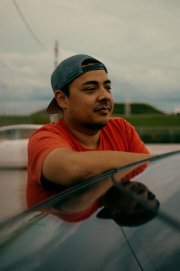 Lenny Sanchez, in a ball cap and orange T-shirt, leans his arm against the back windshield of a car.