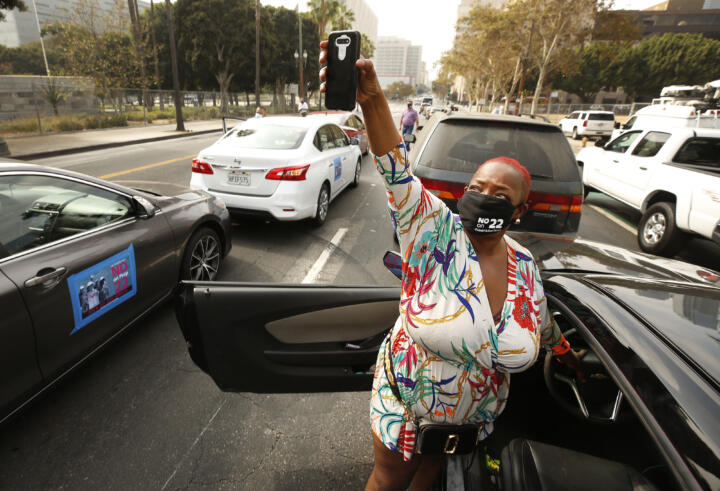 A masked woman, surrounded by parked cars in a demonstration, holds up a cell phone.