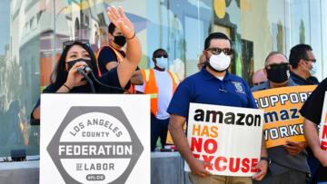 A masked woman stands behind a podium with her hand raised. People next to her are holding signs criticizing Amazon.