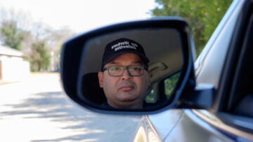 Photograph of a man's reflection in a car's side view mirror. He is wearing a hat that says "Gig Workers Collective"