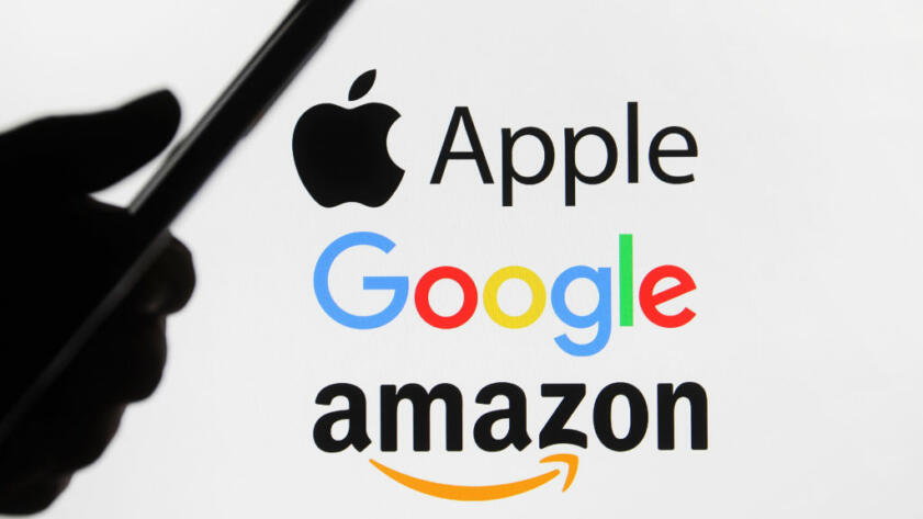 In this photo illustration Google, Amazon and Apple logos are seen behind a silhouette of a hand holding a mobile phone.