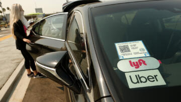 Photo of a car displaying an Uber and Lyft sticker on its window. A passenger is opening the rear door to enter.