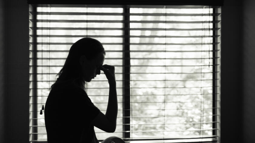 Silhouette of a stressed young woman backlit against a window. She is holding her nose with her hand.