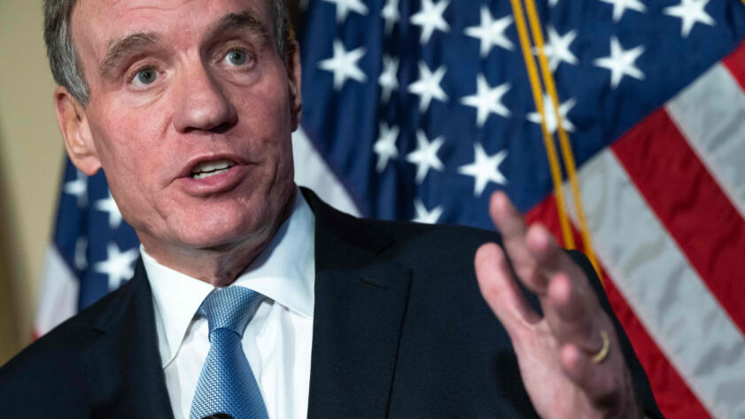 U.S. Sen. Mark Warner standing at a podium with his left hand raised. In the background, there is an American flag.