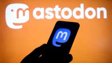 Photo of a silhouette of a hand holding a phone with the Mastodon logo on its screen. The background is the Mastodon logo.