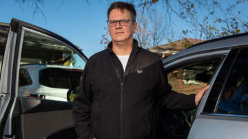 Photograph of Brian Blagoue, a White man with short brown hair. Blagoue is wearing glasses and a black zip-up sweater with the Lyft logo on his upper left chest area. He is standing in front of a silver car with the driver's door open.