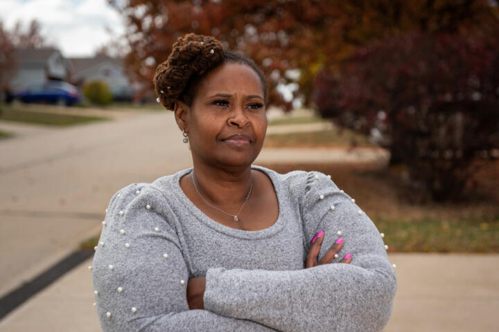 Photograph of Elizabeth Halton, a Black woman. Halton is wearing a gray sweater with white pearls attached around her shoulders. Her arms are crossed and she has a contemplative facial expression.