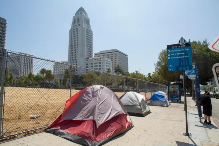 Photograph of three tents lining the sidewalk in Los Angeles. The City Hall building stands on the other side of the block behind the encampments.