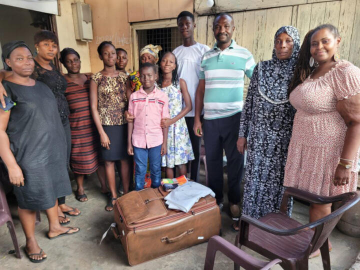 Photograph of Elizabeth Hylton standing with 11 of Elijah Newman's family members. They are standing around a brown suitcase with folded clothes laid on top of it.