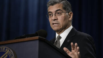 Xavier Becerra stands behind a podium in front of a dark curtained background.