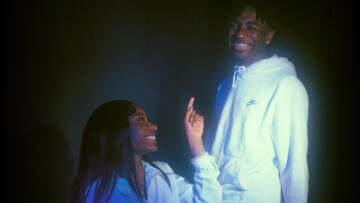 Photo of Mia Townsend and Maurice Newton, two young Black teenagers. Mia, seated and wearing a white collared shirt, is smiling and pointing at Maurice, who is wearing a white Nike hoodie.
