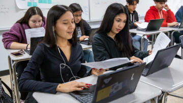 Photograph of 6 high school students on their laptop. Four of the students have long hair, two have short hair. One student has one earbud in their ear.