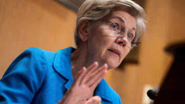 Photograph of Senator Elizabeth Warren at a podium. She is wearing glasses and a blue blazer.
