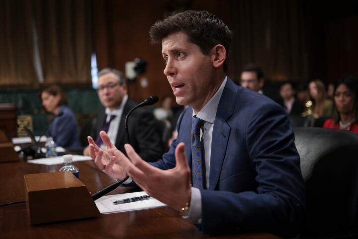 Photograph of Samuel Altman, a man in a suit, sitting at a desk speaking near a microphone.