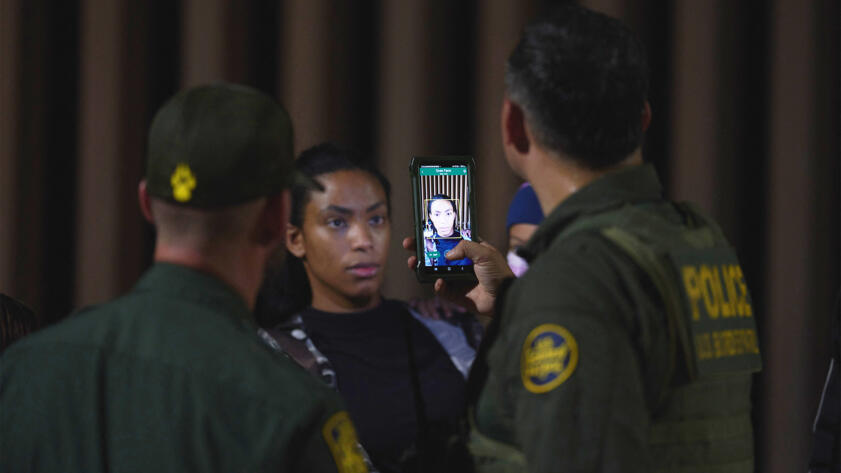 Una mujer, de pie frente a dos hombres, mostrados desde atrás, con uniformes verdes de la Patrulla Fronteriza de Estados Unidos; un agente usa un teléfono inteligente para escanear su rostro.