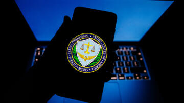 Photograph of a hand holding a phone showing the Federal Trade Commission logo. The hand and phone are backlit by a blue laptop screen, so they just appear as shadows.