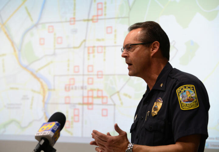 Photograph of Police Chief William Heim standing in front of a microphone and a presentation of a map with various red squares over it.