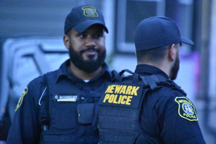 Photograph of two bearded police officers in uniform, both sporting caps and beards. One police officer’s back, whose skin is lighter, is turned towards the camera while the other, whose skin is darker, is facing it.
