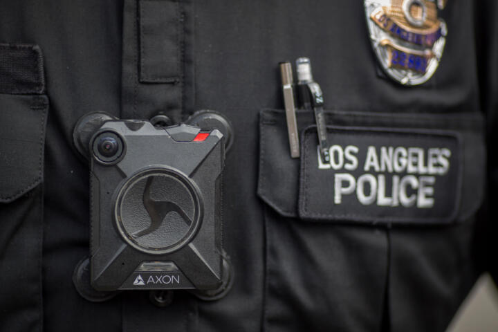 Closeup photograph of a black Axon body camera worn by a police officer. The badge next to it says “LOS ANGELES POLICE.”