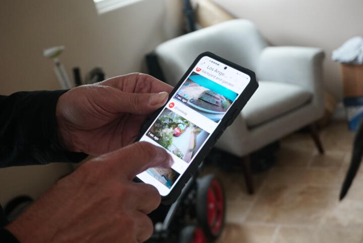 Closeup photograph of Dorini’s hands holding a smartphone. The screen is showing three different camera views on the Ring app. One view is labeled “backyard and garden” and another is labeled “driveway.”