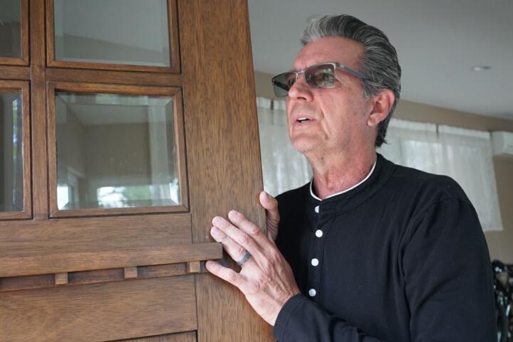 Closeup photograph of Ed Dorini, a White man in his 60s sporting dark gray hair that’s pulled back and tinted sunglasses. Dorini is holding the edge of the door with both hands while looking outside it.
