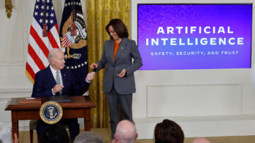 Image of U.S. President Joe Biden seated at a desk, passing a pen to Vice President Kamala Harris, who is standing. The background screen shows the words 'Artificial Intelligence' with the subheading 'Safety, Security, and Trust' displayed prominently.