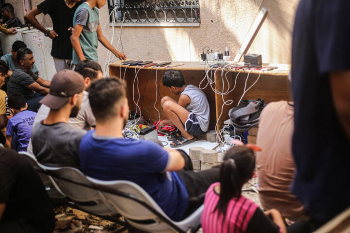 Photo of a Palestinian boy crouched underneath a table with several phones charging on it; he is surrounded by people sitting and waiting with him