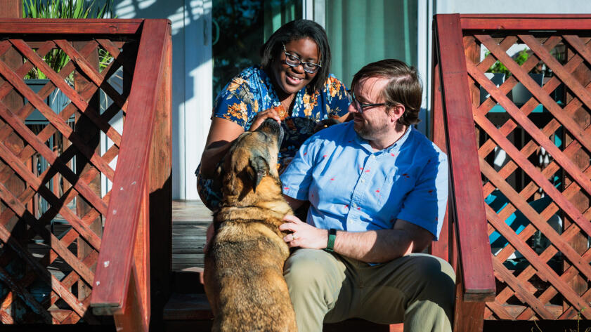 Photograph of Dana, her husband Luke Morris, and their dog Lucy, a German Shepard mix, seated on their back deck