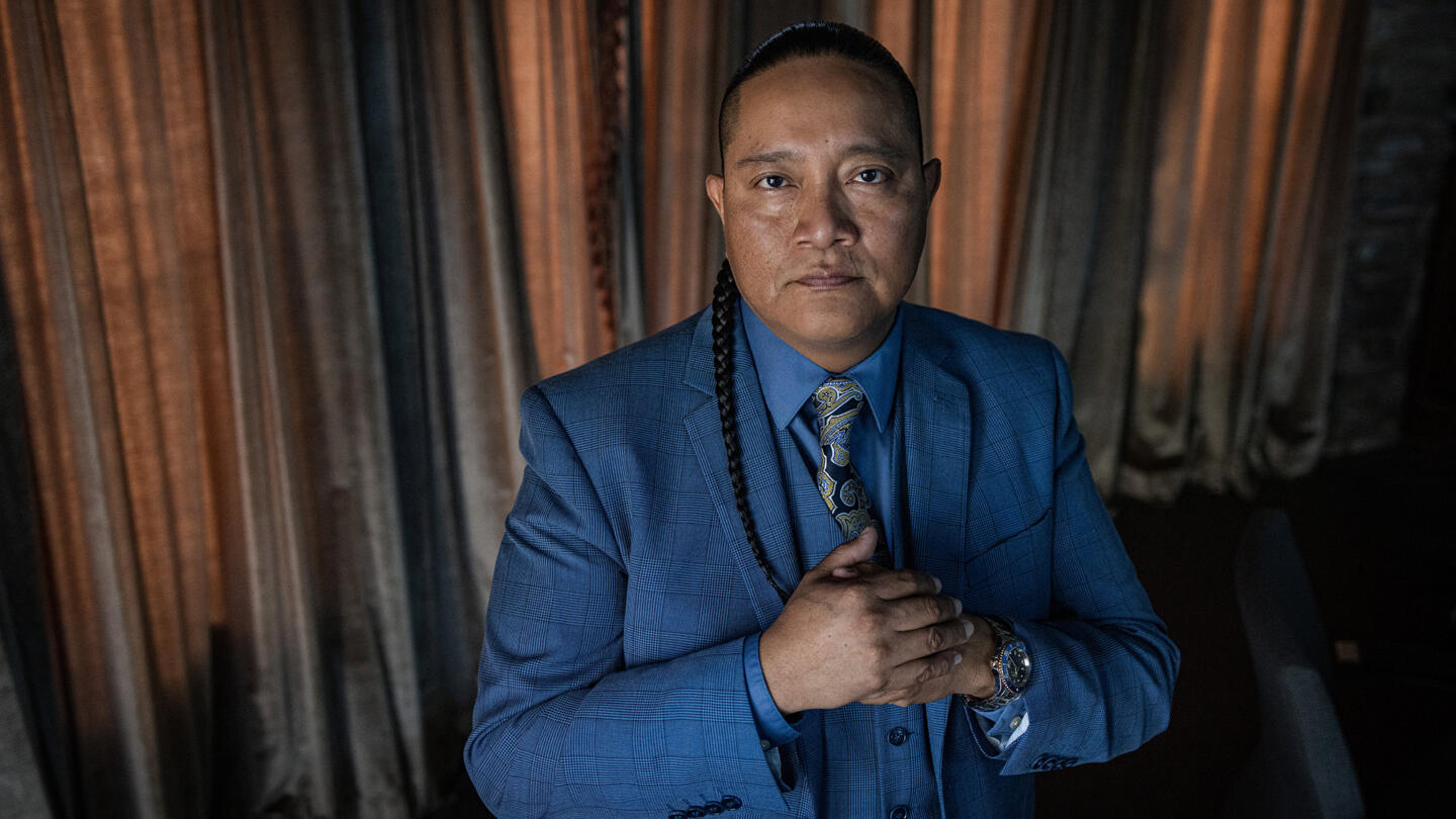 Portrait photograph of Lee Yaiva, an Indigenous man wearing a blue suit and sporting a braided ponytail; his hands are clasped together