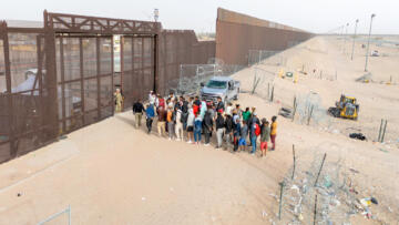 Photograph of panoramic view of immigrants standing in line to cross the U.S.–Mexico border, with one border patrol agent standing in the middle of the gate