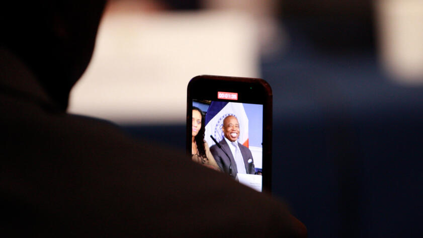 Photograph of Eric Adams smiling, seen through a smartphone recording him