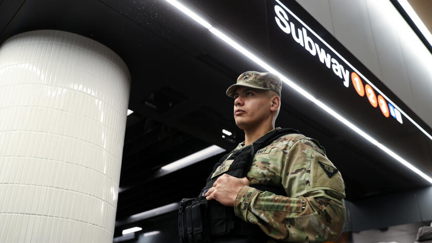 Low-angle photograph of a National Guard soldier in front of a New York City subway sign