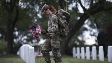 A uniformed soldier with long red hair holding American flags and looking down at a row of tombstones