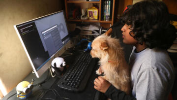 Photo of a young boy seated at his computer, with a small brown dog in his lap