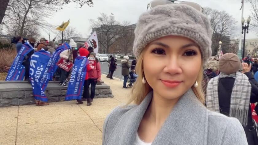 Screenshot of Sonia Ohlala looking to the side of the camera, wearing a gray coat and beanie; behind her is a group of protestors wearing Trump 2020 MAGA flags wrapped around them