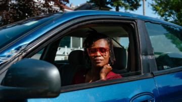 Photo of Melissa Love sitting in the driver’s seat of her blue car, looking straight at the camera