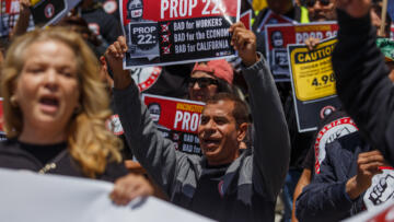  A crowd of protesters hold Prop. 22 signs with their mouths open and fists in the air. One prominent sign reads,  "Unconstitutional Prop 22, Bad for Workers, Bad for the Economy, Bad for California."