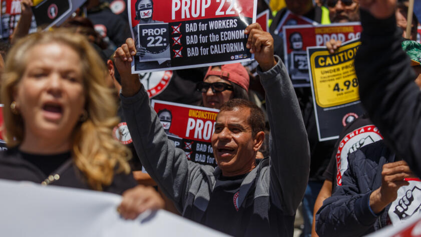 A crowd of protesters hold Prop. 22 signs with their mouths open and fists in the air. One prominent sign reads, "Unconstitutional Prop 22, Bad for Workers, Bad for the Economy, Bad for California."