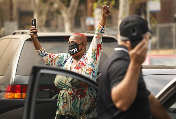 Photograph of a Black woman standing outside her car with her cell phone and arms raised