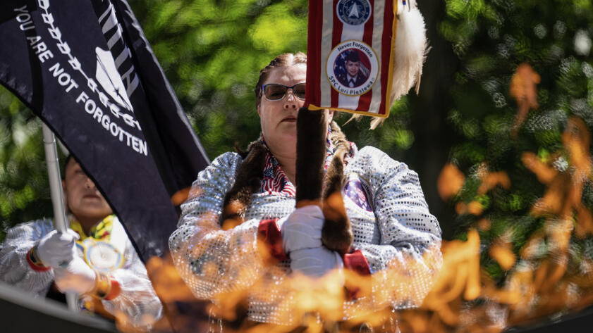 A person in ceremonial attire holds a staff and stands in front of flags during a memorial for Native American war veterans. They are wearing glasses and have braided hair, dressed in a sequined outfit with patches, including one that reads 'Never Forgotten' with an image of a military service member.