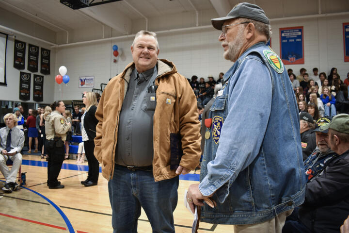 A person wearing jeans, a tucked-in gray shirt and a brown jacket stands next to a person wearing a denim jacket and a gray baseball cap. The jacket has patches related to American wars. The setting is a high school gymnasium.