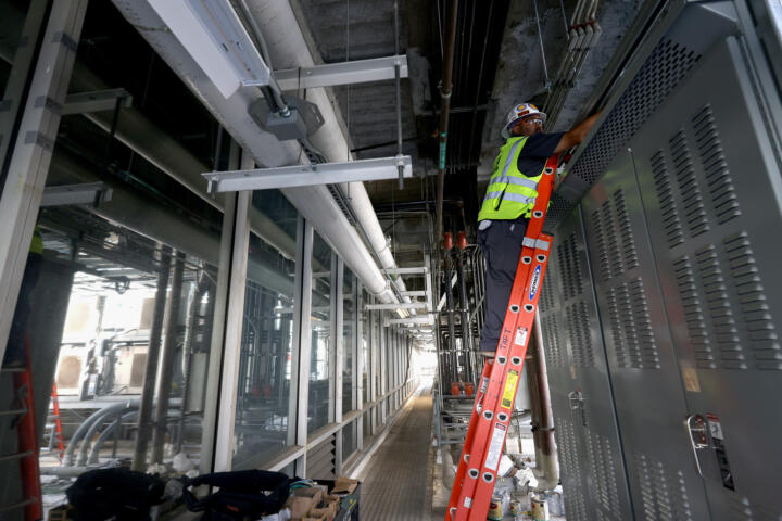 A worker wearing a high-visibility vest, hard hat, and safety glasses stands on an orange ladder, installing or maintaining electrical or mechanical equipment inside an industrial facility. The setting is a long, narrow corridor lined with glass windows on one side and metal cabinets with ventilation panels on the other, with exposed pipes and ducts running along the ceiling.