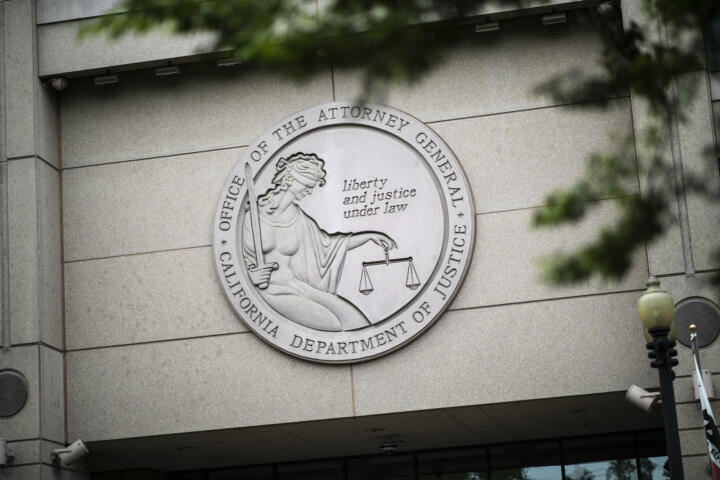 A large circular emblem on the exterior of a stone building displays the seal of the California Department of Justice, Office of the Attorney General. The emblem features an image of a blindfolded figure holding a set of scales and a sword, with the inscription "Liberty and Justice Under Law." Green tree branches partially frame the top of the image, and a lamppost stands in the foreground. Security cameras are mounted on the building's facade.