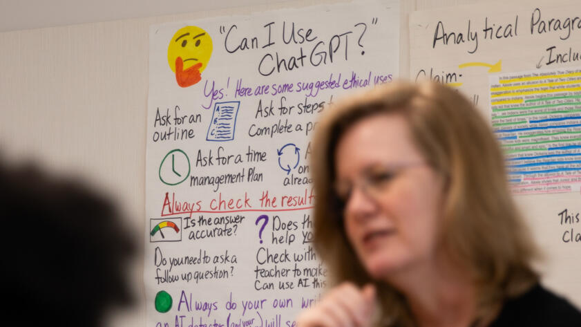 A poster hung up on the wall of a high school classroom with information on how students can use ChatGPT, an AI platform, for schoolwork. The poster is titled: "Can I use ChatGPT?" next to a thinking emoji. English teacher Jen Roberts can be seen in the foreground of the frame.