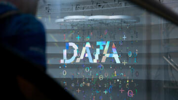 A large, illuminated sign spelling "DATA" is suspended from the ceiling, with holographic numbers, plus signs, and other symbols hanging around it. The reflective installation casts colorful light patterns. In the foreground, a blurred figure wearing a blue jacket is visible, partially obstructed by a glass surface. The scene takes place in a modern conference or exhibition space with a high ceiling and metal framework.