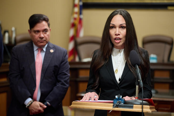 A person, with long black hair and wearing a black blazer with a white blouse, speaks while standing behind a wooden lectern with a microphone. A lawmaker wearing a gray suit and pink tie can be seen in the background on the left side of the frame.
