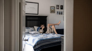 A young boy lounges on a bed with a blue and white patterned blanket, holding a camera, in a softly lit bedroom decorated with family photos and a framed sign that reads "It's always better when we're together."