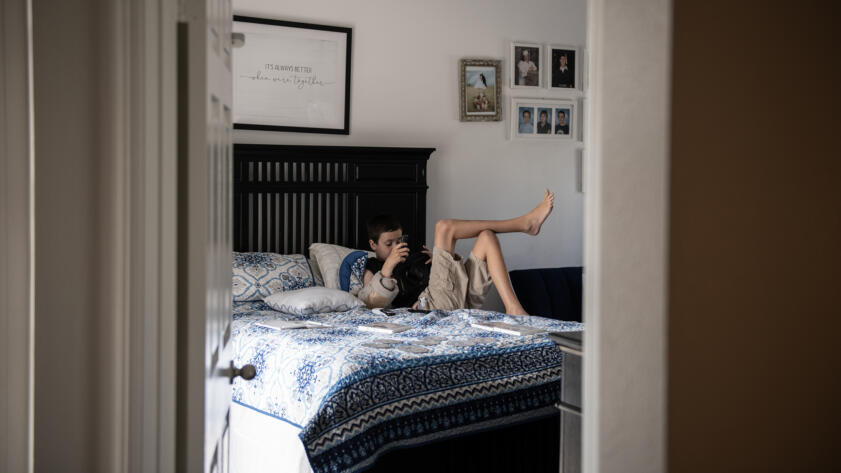 A young boy lounges on a bed with a blue and white patterned blanket, holding a camera, in a softly lit bedroom decorated with family photos and a framed sign that reads "It's always better when we're together."