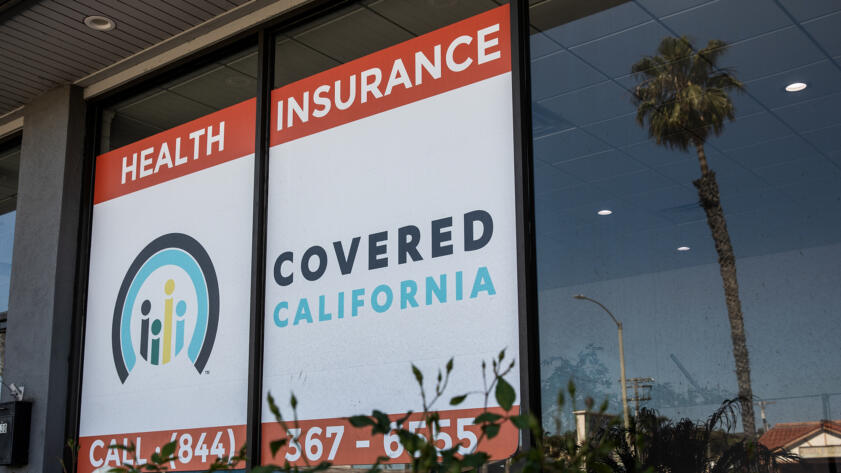 A storefront window displays a large sign reading “HEALTH INSURANCE” and “COVERED CALIFORNIA” with the organization’s logo and a partial phone number. Reflections of a palm tree and a streetlight are visible in the glass.